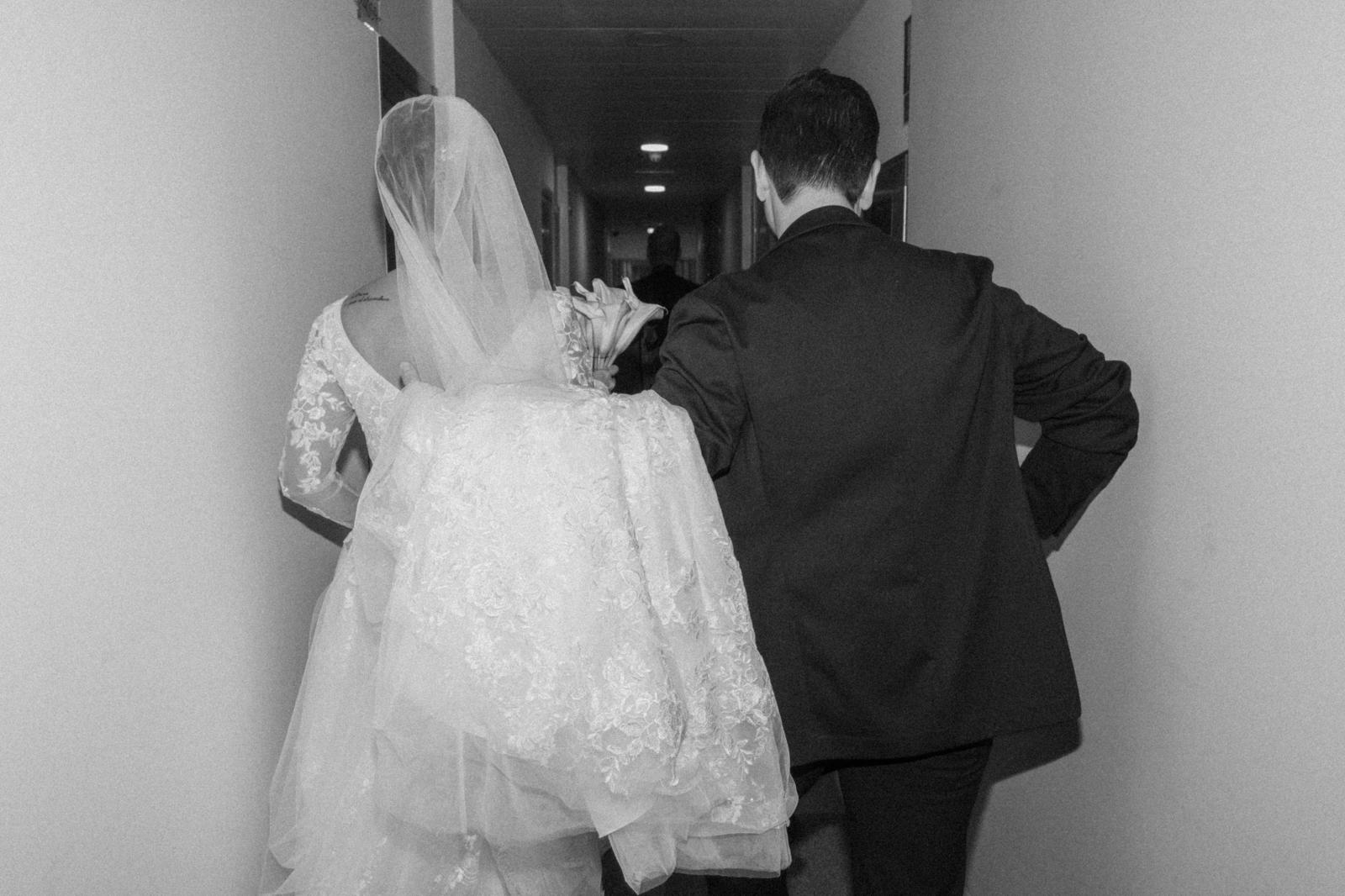 Novia de encaje vista de espaldas con un ramo de callas, caminando por un pasillo, en blanco y negro